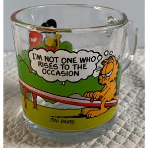 May include: Clear glass mug featuring Garfield with the quote "I'm not one who rises to the occasion." The mug has a handle and a cartoon scene with green, red, and yellow colors. The artist's signature, Jim Davis, is at the bottom.