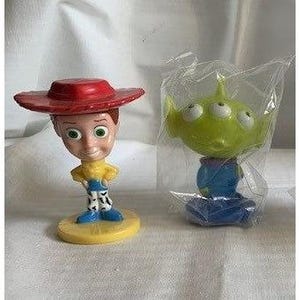 May include: Two Toy Story character figurines. One is Woody, with a red hat, yellow shirt, blue pants, and a yellow base. The other is an Alien, green with three eyes, in a plastic bag, on a blue base.