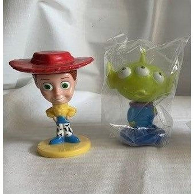 May include: Two Toy Story character figurines. One is Woody, with a red hat, yellow shirt, blue pants, and a yellow base. The other is an Alien, green with three eyes, in a plastic bag, on a blue base.