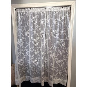 May include: A white lace curtain panel featuring a repeating floral pattern with roses and scrolling vines. The sheer fabric has a delicate mesh texture, a gathered top header, and a solid lace border along the bottom edge, hanging in a doorway.