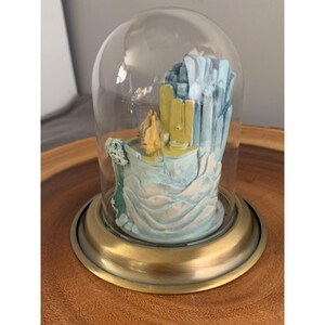 May include: A glass dome with a brass base encloses a miniature ceramic sculpture of a building with blue and yellow accents. The sculpture is set on a base that resembles a wave.