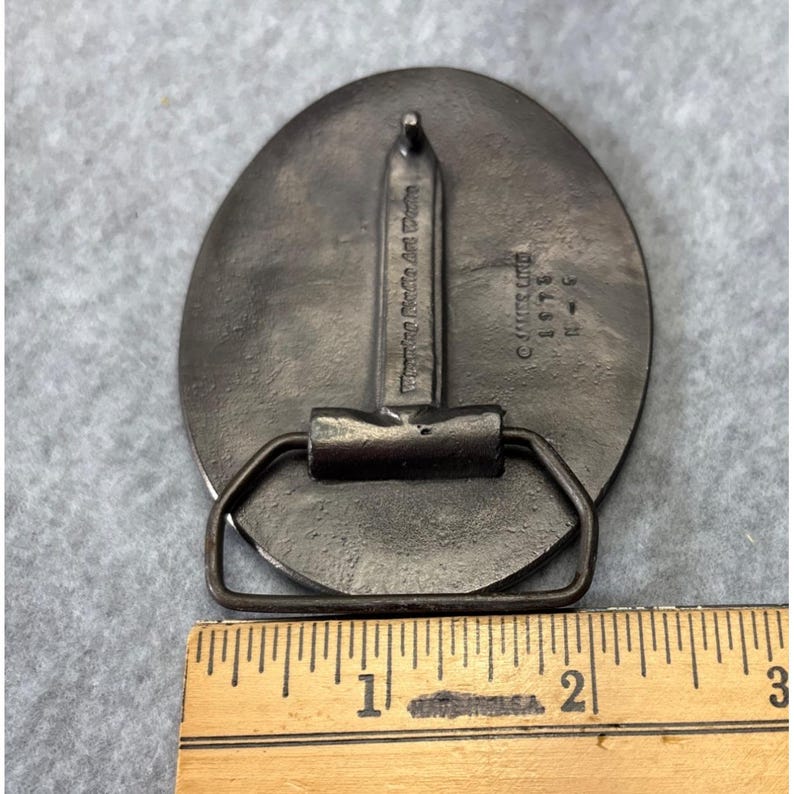 May include: A vintage metal belt buckle with an oval design. The buckle has a rectangular bar and a loop for the belt. The text "Wyoming Rodeo Art Works" and "&copy; James Lino 1975" are visible. The buckle measures approximately 2.5 inches in length.
