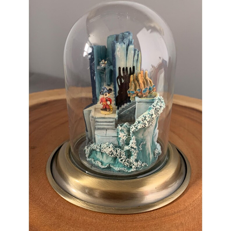 May include: A glass dome with a brass base encloses a miniature scene featuring Mickey Mouse standing on a staircase in front of a blue and white building. The scene is set against a backdrop of blue water with white foam.