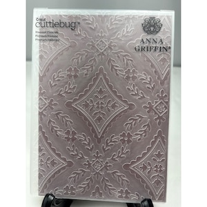 May include: A rectangular Cricut Cuttlebug embossing folder by Anna Griffin featuring a Diamond Flourish pattern. The clear plastic folder displays a mauve-colored insert with a repeating white floral and diamond lattice design throughout.