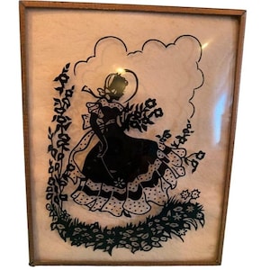 May include: A framed silhouette art piece featuring a black figure of a woman holding flowers. The silhouette is set against a light background, framed in a wooden frame. The woman is wearing a dress with polka dots and a hat.
