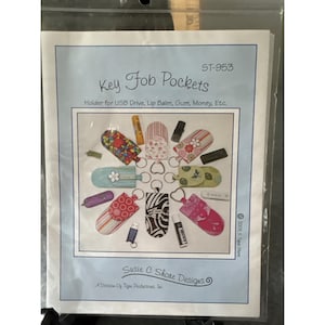 May include: A package for "Key Fob Pockets" with various designs. The text reads "Holder for USB Drive, Lip Balm, Gum, Money, Etc." and "Susie C Shore Designs". The image shows examples of key fob pockets.