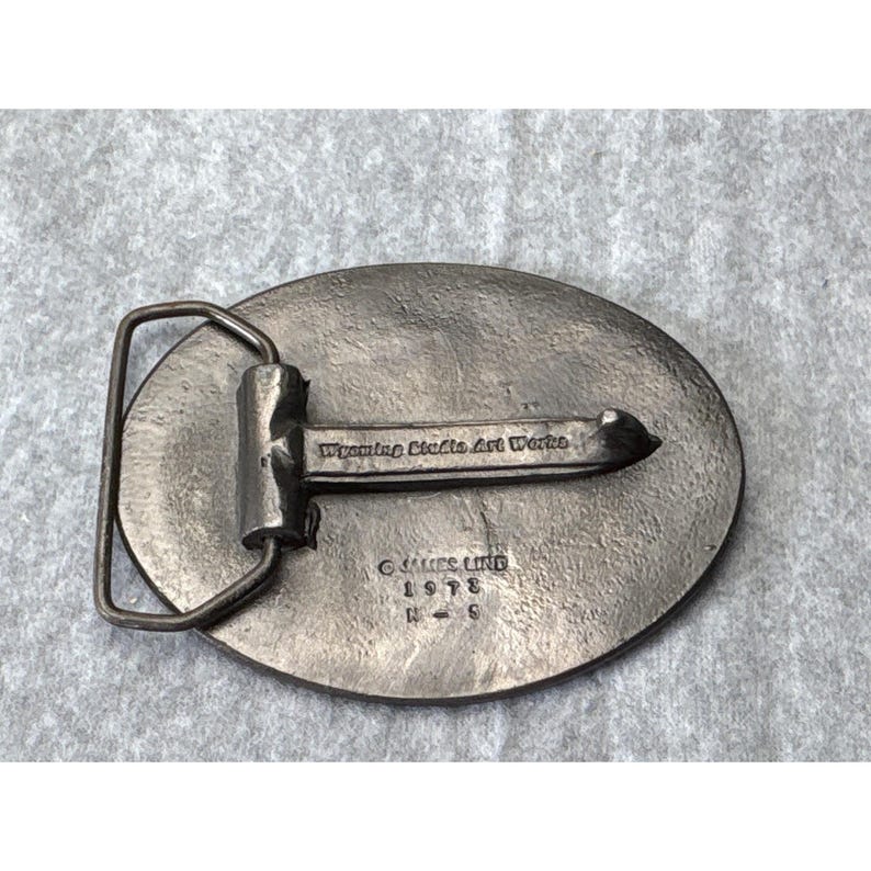 May include: Vintage metal belt buckle with an oval shape and a silver finish. The buckle features a rectangular bar and a prong for securing the belt. The text "Wyoming Studio Art Works" is visible, along with the name "JAMES LIND" and the year 1978.