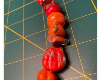 Può includere: Un ciondolo fatto a mano con perline di vetro arancioni e rosse disposte verticalmente. Le perline hanno varie forme, tra cui rotonde e a forma di zucca, con dettagli in filo d'argento. Il ciondolo è posto su un tappetino da taglio verde.