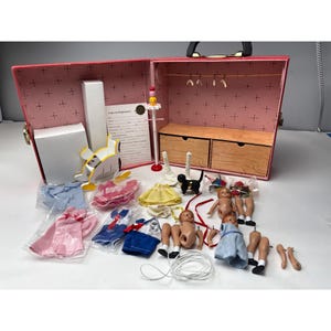 May include: A vintage doll wardrobe set, open to reveal a pink interior with a clothes rack and drawers. The set includes doll clothes, doll parts, and accessories. The wardrobe is red with a black handle.