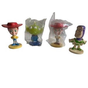 May include: Four Toy Story character figurines. Woody, with a red hat and yellow shirt, stands on a yellow base. An alien with three eyes is in a plastic bag. Jessie, with a red hat, is in a plastic bag. Buzz Lightyear stands on a base.