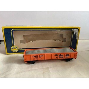 Puede incluir: Un vagón de tren modelo naranja con el texto "D&RGW 561 63" y "Rio Grande" en el lateral. El vagón está lleno de material gris y se encuentra frente a su caja amarilla y azul.