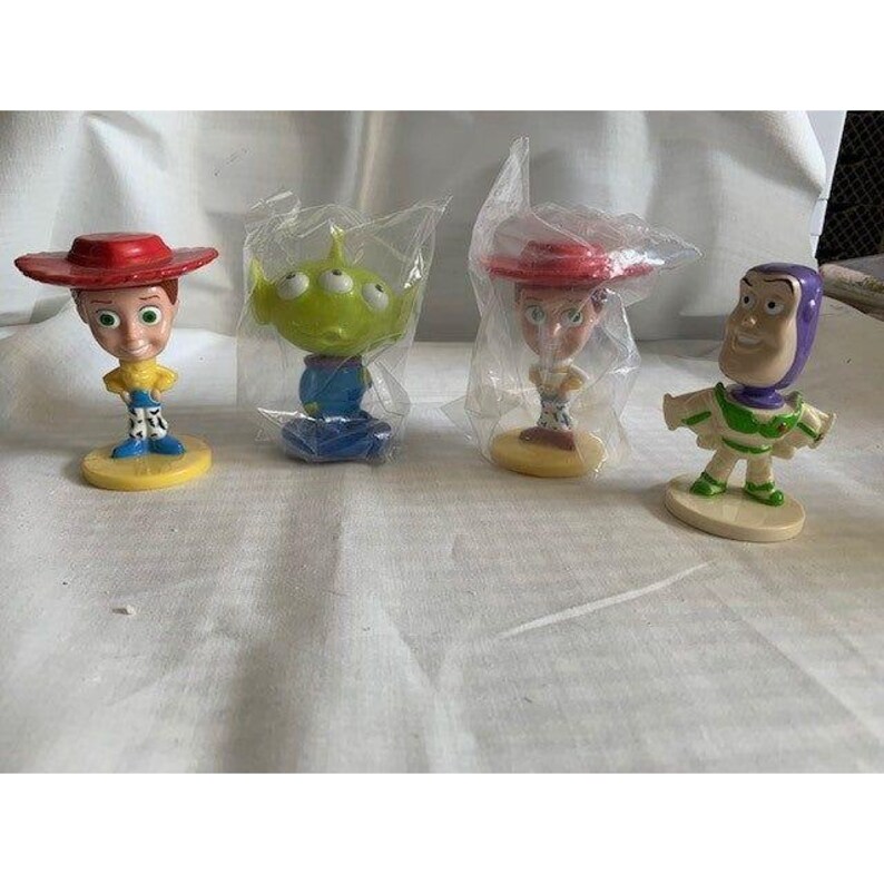 May include: Four collectible figurines from the animated film series. The figures include Woody, an alien, Jessie, and Buzz Lightyear. Each figure is on a small base. The Woody and Jessie figures have red hats. The alien has three eyes.
