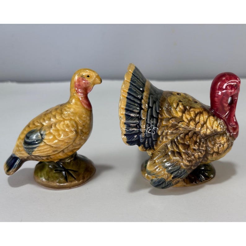 May include: A pair of ceramic turkey figurines, likely salt and pepper shakers. The turkeys are painted in shades of yellow, brown, and blue, with red accents on their heads. One turkey faces left, the other faces right.