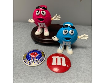 以下が含まれることがあります： ピンクとブルーの2体のM&M'sキャラクターが木製の台座の上に立っています。ピンクのM&M'sキャラクターは白い靴を履いており、ブルーのM&M'sキャラクターは白い靴を履いています。赤いM&M'sロゴと、「Celebrating 75 Years of the New Millennium」という文字が書かれた丸い白地に金色の縁取りのステッカーがあります。