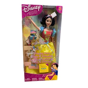 May include: A Disney Snow White doll in a box. The doll wears a yellow dress with a red and sequined bodice and blue sleeves. The box reads "Princess Party" in multiple languages and features images of Snow White and the Seven Dwarfs.