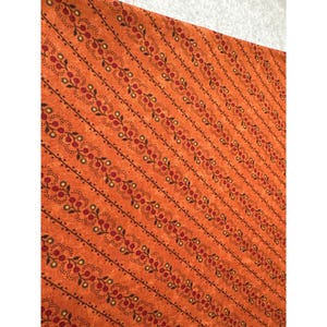 May include: Orange fabric with a repeating pattern of diagonal lines. Each line features a series of small red and yellow berries with green leaves, connected by a dotted line. The fabric has a textured appearance.