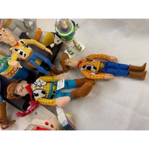 May include: A collection of Toy Story character plush dolls, including Woody and Buzz Lightyear. Woody dolls wear a yellow shirt with black cow spots, blue pants, and a brown cowboy hat. Buzz Lightyear is in a green and white spacesuit.