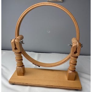 May include: A light-colored wooden embroidery hoop stand featuring a circular frame mounted on two vertical, turned-wood supports. The base is a rectangular wooden plank, and the hoop is secured by metal hardware on both sides, allowing for rotation.
