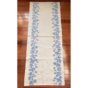 May include: A rectangular, off-white table runner with a repeating pattern of blue leaves and vines along the edges. The runner is made of a light-colored fabric and is displayed on a wooden surface.