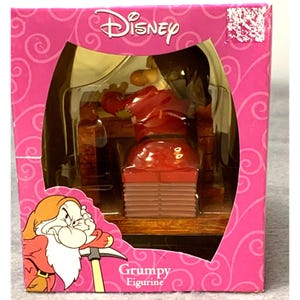 May include: A Disney Grumpy figurine packaged in a bright pink box with swirling patterns. The box features an illustration of the character Grumpy and the text Grumpy Figurine. The figurine inside is visible through a clear plastic window.