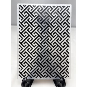 Puede incluir: Una carpeta de relieve rectangular con una construcción de plástico transparente y un patrón geométrico negro en relieve. El diseño consiste en un intrincado motivo de llave griega repetitivo que cubre toda la superficie de la herramienta.