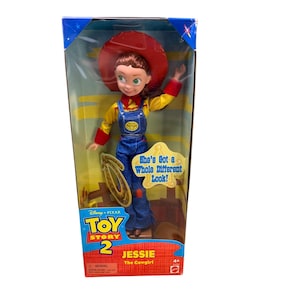 Może przedstawiać: Plastikowa lalka Jessie z Toy Story 2 w pudełku. Ma na sobie czerwony kapelusz, ż&oacute;łtą koszulę, niebieskie ogrodniczki i brązowe buty. Pudełko jest ż&oacute;łte i niebieskie z logo Toy Story 2 i tekstem "She's Got a Whole Different Look!" i "Jessie The Cowgirl".