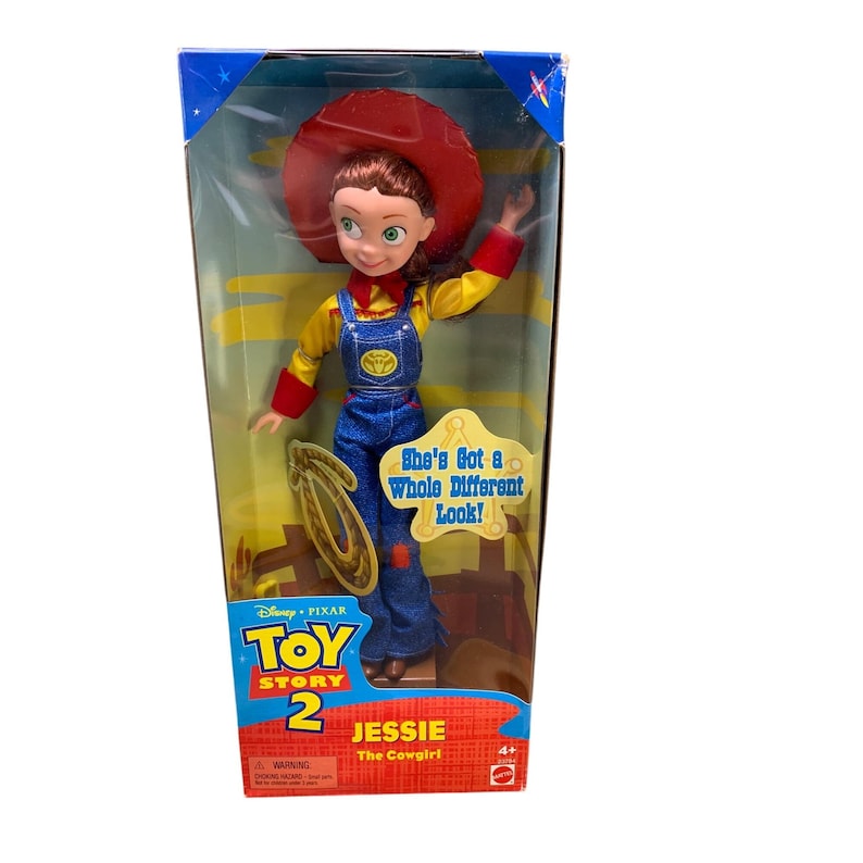 Może przedstawiać: Plastikowa lalka Jessie z Toy Story 2 w pudełku. Ma na sobie czerwony kapelusz, ż&oacute;łtą koszulę, niebieskie ogrodniczki i brązowe buty. Pudełko jest ż&oacute;łte i niebieskie z logo Toy Story 2 i tekstem "She's Got a Whole Different Look!" i "Jessie The Cowgirl".