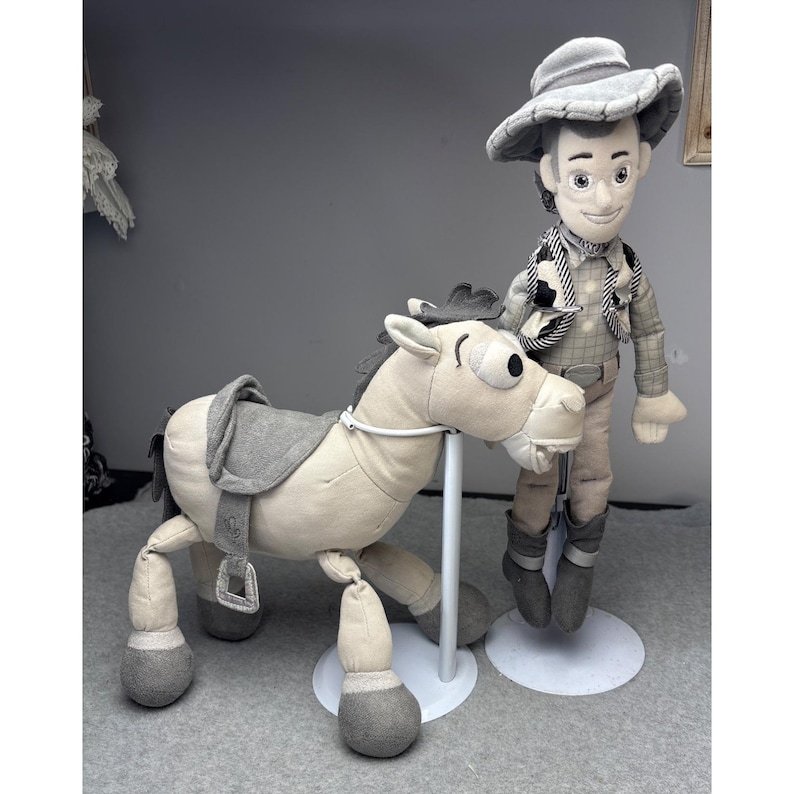 K&ouml;nnte beinhalten: Ein Pl&uuml;schtiersatz mit einer Cowboy-Puppe und einem Pferd. Die Cowboy-Puppe tr&auml;gt einen Hut, eine Weste und Stiefel. Das Pferd hat einen Sattel und einen Zaum. Beide Spielzeuge sind haupts&auml;chlich beige und grau, mit wei&szlig;en Akzenten. Sie stehen auf wei&szlig;en St&auml;ndern.