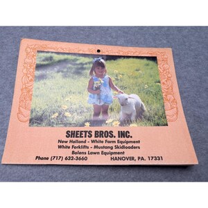 May include: A light brown framed photograph showing a young girl in a blue dress with a white lamb in a field. The frame has decorative edges. The text "SHEETS BROS. INC." and contact details are printed at the bottom of the frame.