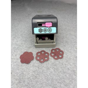 May include: A gray and pink flower-shaped paper punch with three cut-out flower shapes. The punch has a pink slider and a light blue label with flower graphics. The cut-out flowers are a reddish-brown color.