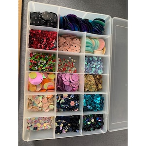 May include: A clear plastic organizer filled with various colors of sequins. The sequins are in different shapes and sizes, including circles and flower shapes. Colors include black, red, pink, green, gold, and teal.