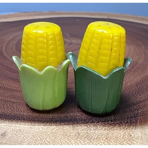 May include: Two yellow corn on the cob salt and pepper shakers with green husks. The shakers are made of plastic and have a realistic corn cob design.