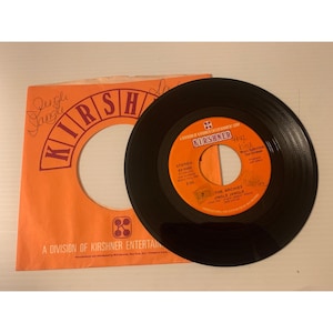 May include: A vintage vinyl record and its orange sleeve. The record is black with orange and white labels, featuring the text "The Archies" and "Jingle Jangle." The sleeve has the Kirshner logo and the words "Jungle Jangle."