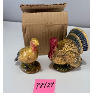 May include: A pair of ceramic turkey figurines, one standing and one sitting, with yellow, brown, red, and blue coloring. The figurines are on green bases. A cardboard box is in the background. A pink sticker with the text "P8427" is in the foreground.