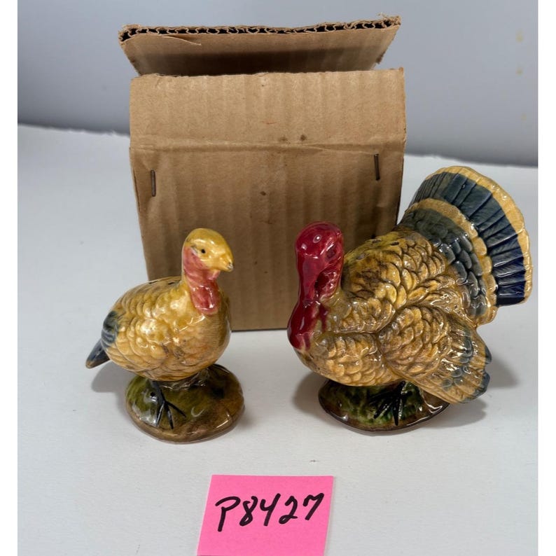 May include: A pair of ceramic turkey figurines, one standing and one sitting, with yellow, brown, red, and blue coloring. The figurines are on green bases. A cardboard box is in the background. A pink sticker with the text "P8427" is in the foreground.