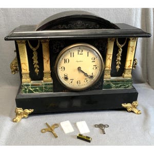 May include: Antique black mantel clock with a cream-colored clock face and gold accents. The clock features decorative columns, ornate detailing, and a curved top. Includes clock keys and a level.