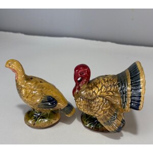 May include: A pair of ceramic turkey figurines, likely salt and pepper shakers. One turkey is standing, the other is fanned out. The turkeys are primarily tan with red, blue, and green accents. They are on a green base.