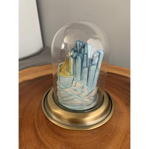 May include: A miniature glass dome with a gold base encloses a blue and yellow ceramic sculpture of a city skyline. The sculpture is on a wooden surface.