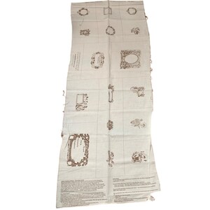 May include: An off-white fabric with a grid pattern and brown decorative elements. The fabric features framed designs, floral motifs, and text blocks. The edges of the fabric are unfinished.