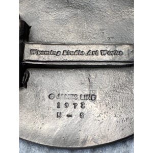 May include: A close-up of a silver-toned metal object with the text "Wyoming Studio Art Works." Below the text are the words "&copy; JAMES LIND 1973 N = 5."