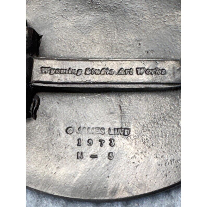 May include: A close-up of a silver-toned metal object with the text "Wyoming Studio Art Works." Below the text are the words "&copy; JAMES LIND 1973 N = 5."