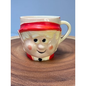 May include: A vintage plastic mug shaped like an elf's face. The elf has a red hat, blonde hair, and a smiling face.