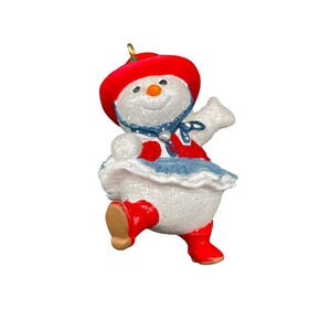 May include: A whimsical snowman ornament with a red hat, vest, and boots, and a blue scarf and skirt. The snowman has a carrot nose and a cheerful expression. The ornament is designed with glittery accents, adding a festive touch to holiday decor.