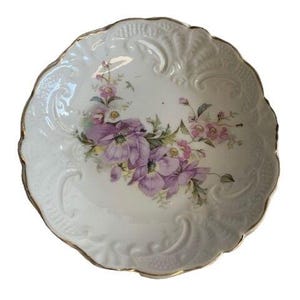 May include: White porcelain decorative plate with a scalloped edge and gold trim. The plate features a floral design with purple and pink flowers and green leaves. The plate has a textured surface.