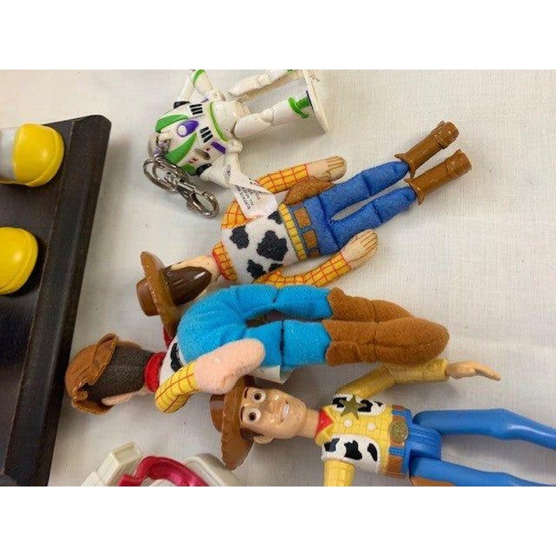 May include: Several Toy Story character figurines, including Woody and Buzz Lightyear, are arranged on a white surface. Woody figures wear cowboy hats, vests, and blue pants. Buzz Lightyear is in a white and green spacesuit.