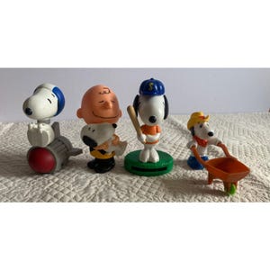 May include: Four Peanuts character figurines. Snoopy in a baseball uniform, Snoopy in a cowboy hat pushing a wheelbarrow, Snoopy on a rocket, and Charlie Brown holding Snoopy. The figurines are on a white surface.