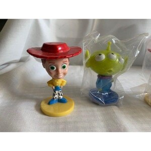 May include: Two Toy Story character figurines. One is a Jessie figurine with a red hat, yellow shirt, and blue pants. The other is a green alien figurine in a plastic bag. Both figurines are on a white surface.