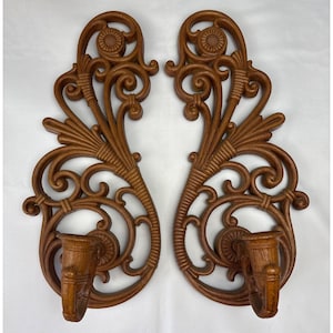 Könnte beinhalten: Paar verzierter, brauner Wandleuchter. Jeder Leuchter hat einen Kerzenhalter und aufwendige Schnörkel mit floralen und Blattdetails. Die Leuchter bestehen aus einem holzähnlichen Material und sind für die Wandmontage konzipiert.