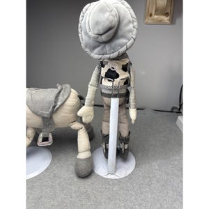 K&ouml;nnte beinhalten: Eine Cowboy-Puppe aus Pl&uuml;sch mit grauem Hut, Weste und Stiefeln steht neben einem Pl&uuml;schpferd mit Sattel. Die Cowboy-Puppe hat eine schwarz-wei&szlig;e Kuhfellweste und steht auf einem wei&szlig;en St&auml;nder. Das Pferd ist hellbeige mit einem grauen Sattel.