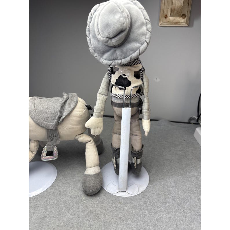 K&ouml;nnte beinhalten: Eine Cowboy-Puppe aus Pl&uuml;sch mit grauem Hut, Weste und Stiefeln steht neben einem Pl&uuml;schpferd mit Sattel. Die Cowboy-Puppe hat eine schwarz-wei&szlig;e Kuhfellweste und steht auf einem wei&szlig;en St&auml;nder. Das Pferd ist hellbeige mit einem grauen Sattel.